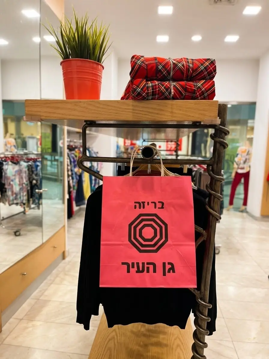 שקית נייר ורודה של בריזה תלויה על מתלה ברזל מול חולצה שחורה, מעליה מדף עץ עם עציץ וחולצות משבצות מקופלות