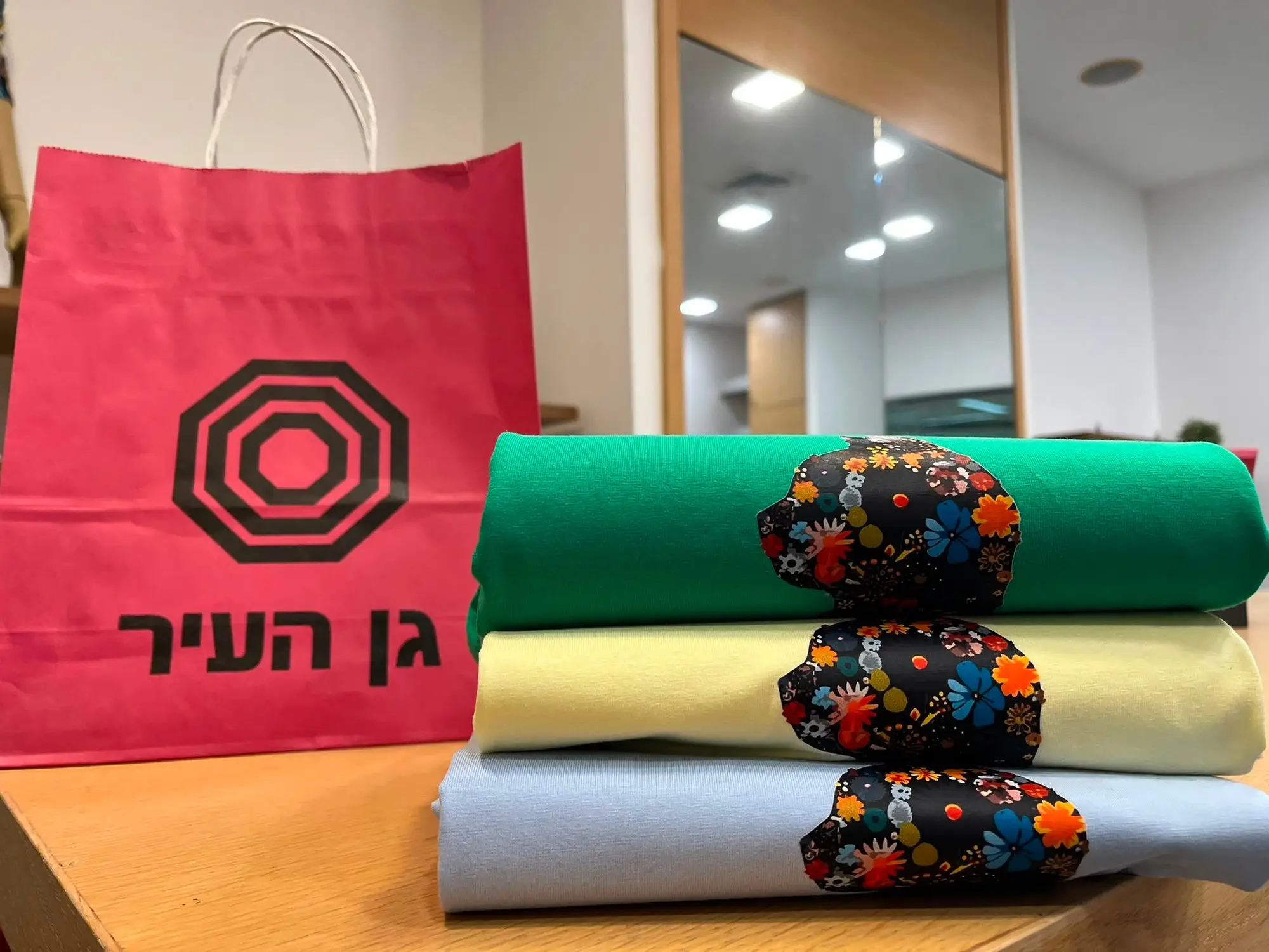 שלוש חולצות מקופלות בירוק, צהוב ותכלת עם הדפס פרחוני, לצד שקית נייר אדומה של גן העיר